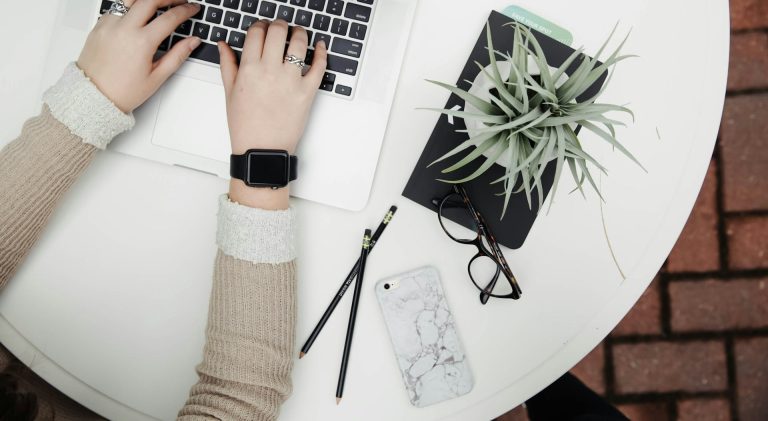 Mains tapant sur un clavier près d'un carnet, de lunettes et d'une plante sur une table.